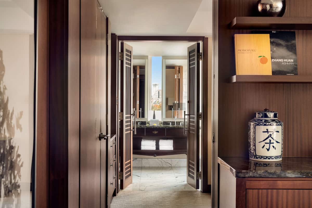 A hallway with wooden doors leading to a bathroom with a marble floor and a sink. There are books on a shelf and a decorative vase on a counter.