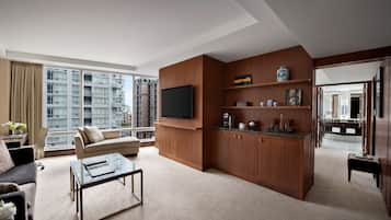 A modern living room with a sofa, coffee table, and TV. It features wooden paneling, a large window with a city view, and a kitchen area with cabinets and a countertop.