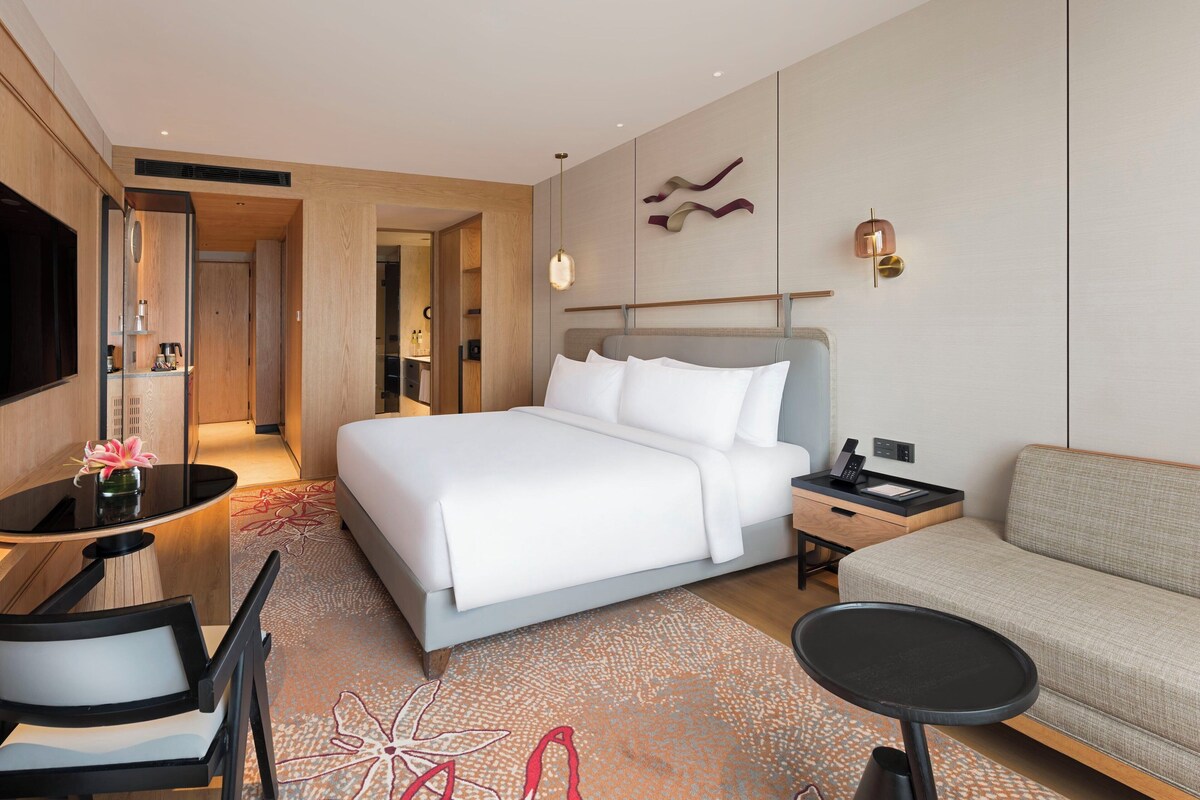 A modern hotel room with a large bed, a sofa, a desk with a chair, and a TV.