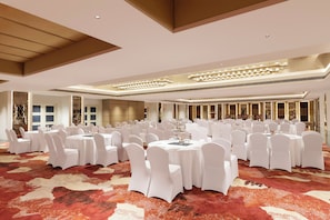 Un salón de banquetes con mesas redondas y sillas blancas, alfombra estampada y un diseño de techo moderno.
