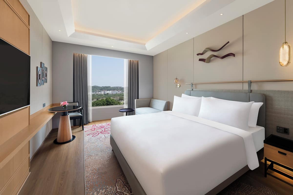 A modern hotel room with a large bed, a TV, a dining area, and a view of the city.