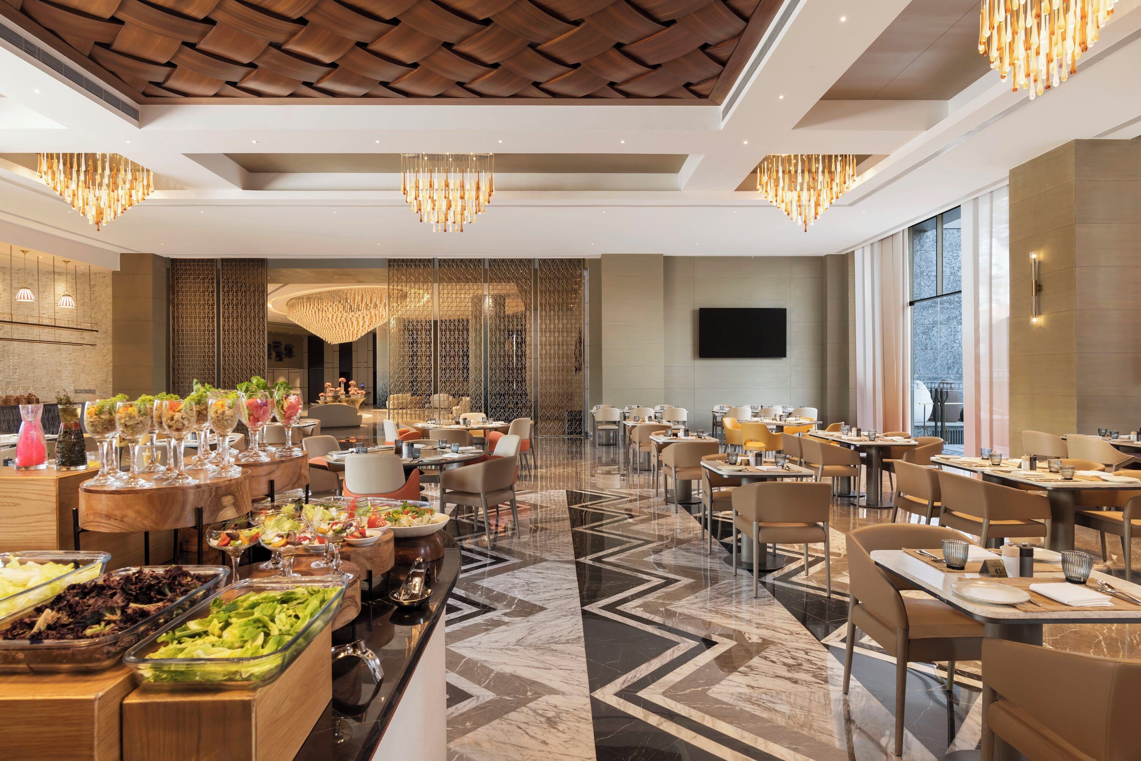 Un amplio comedor de hotel con varias mesas, un bufé con diversas opciones de comida y un moderno candelabro.