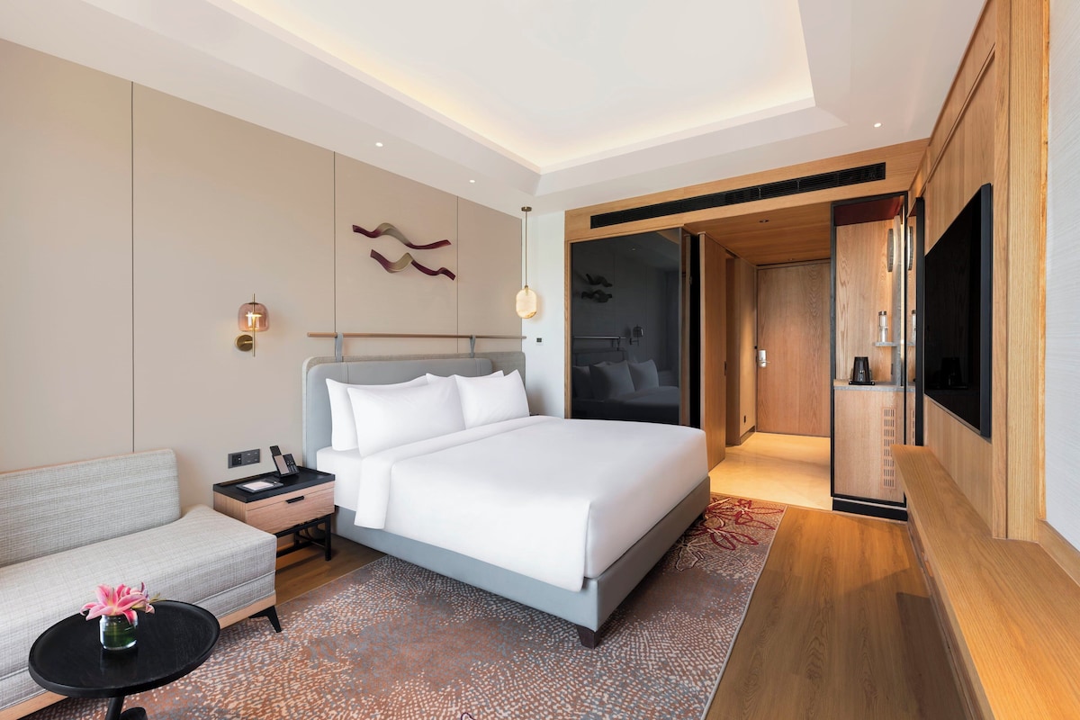 A modern hotel room with a large bed, a sofa, a small table with a vase, a TV, and a sliding door leading to a bathroom.