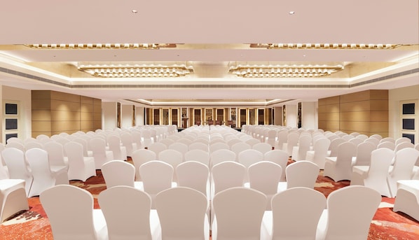 Un amplio salón de conferencias con filas de sillas blancas, un escenario con un atril y una moderna lámpara de araña.
