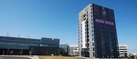 A modern hotel building with a 'NOW OPEN' sign, surrounded by other commercial buildings.