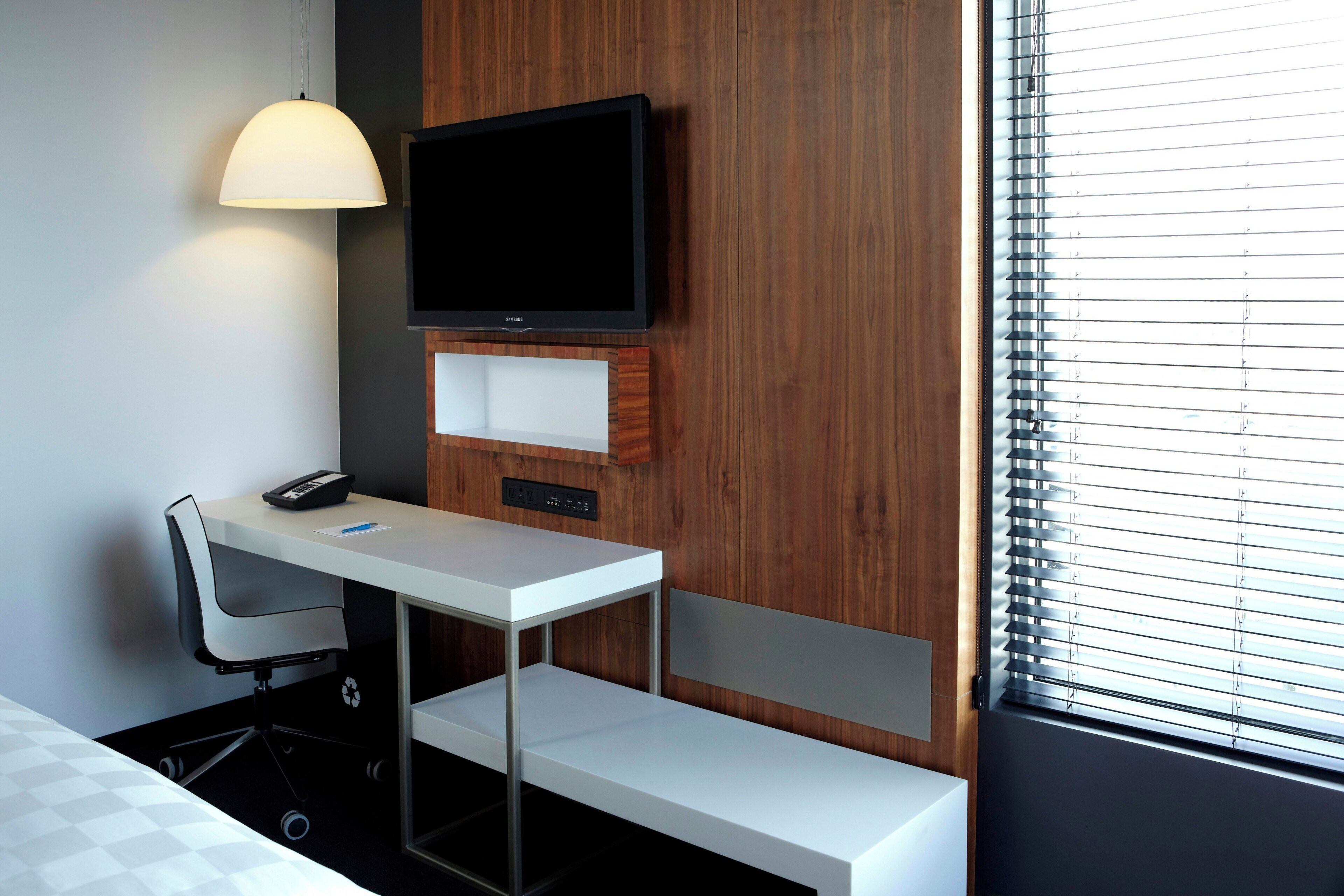 Habitación de hotel con escritorio, silla, televisor y ventana con persianas.