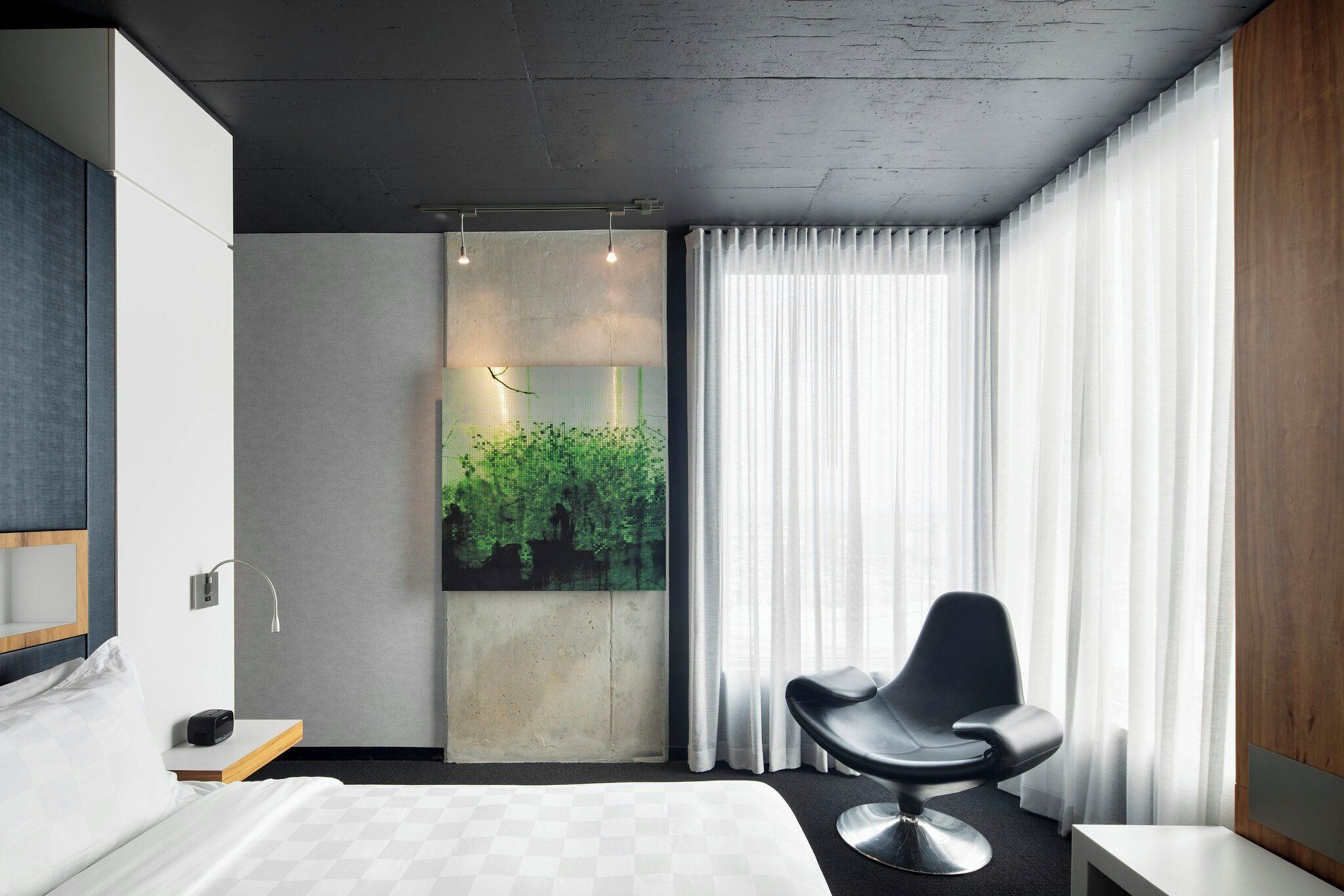 Una habitación de hotel moderna con una cama, un escritorio, una silla, un ventanal con cortinas y una obra de arte colgada en la pared.