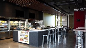 Um café moderno com um balcão central, banquetas de bar e diversas opções de comida e bebida.