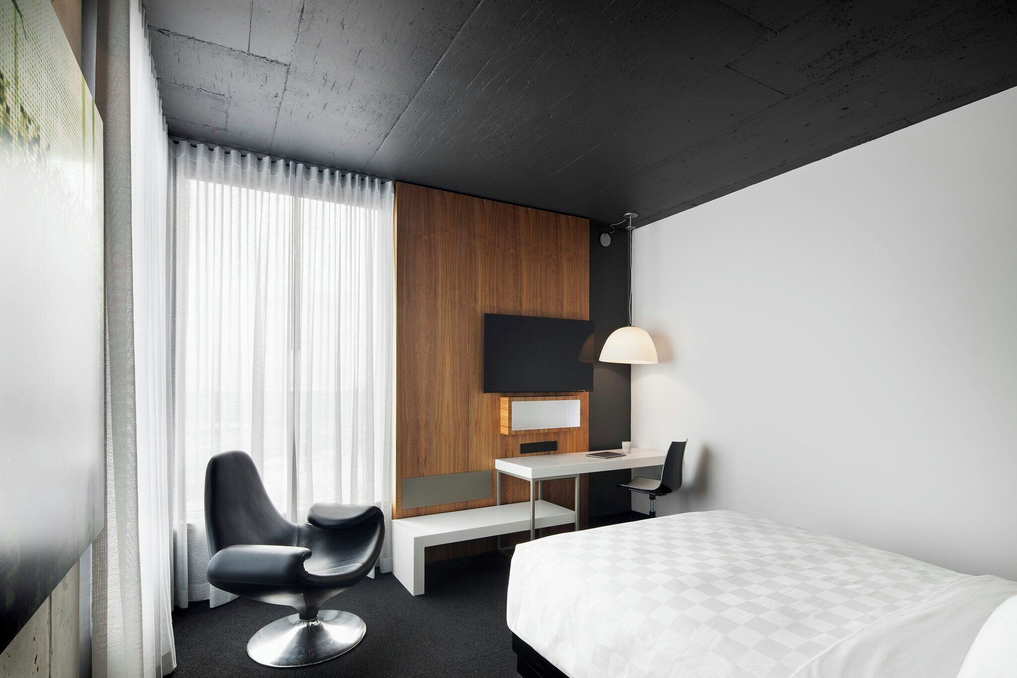 Quarto de hotel com uma cama, escrivaninha, cadeira, luminária e uma janela grande com cortinas.