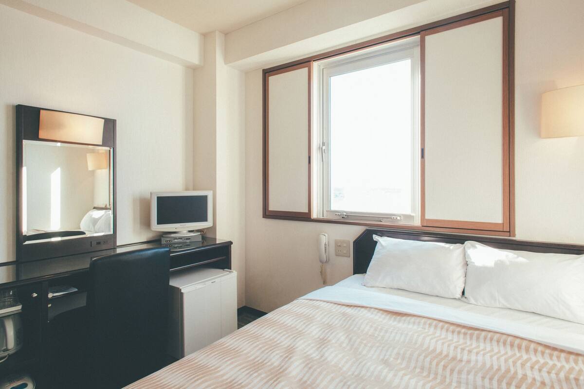 Una camera d'albergo con un letto, una scrivania con un computer, una sedia, un telefono e una finestra.