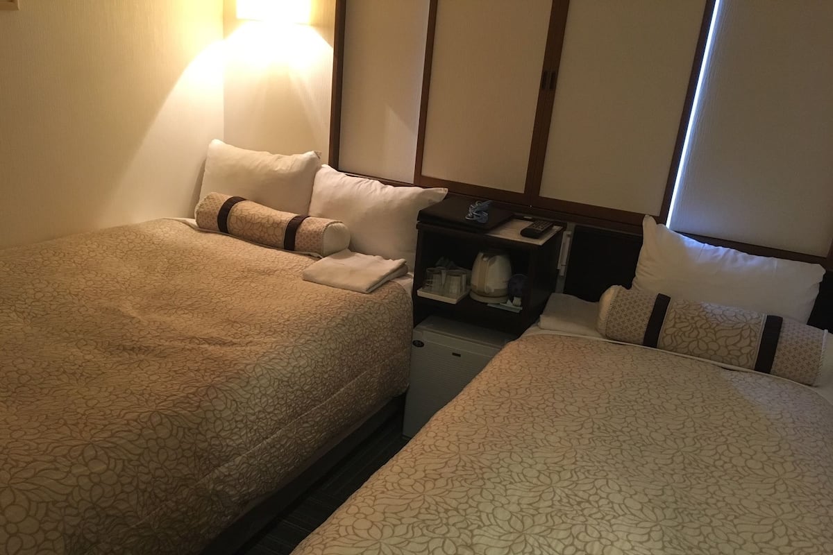 Camera d'albergo con due letti, un comodino con una lampada e un tavolino con un telecomando.