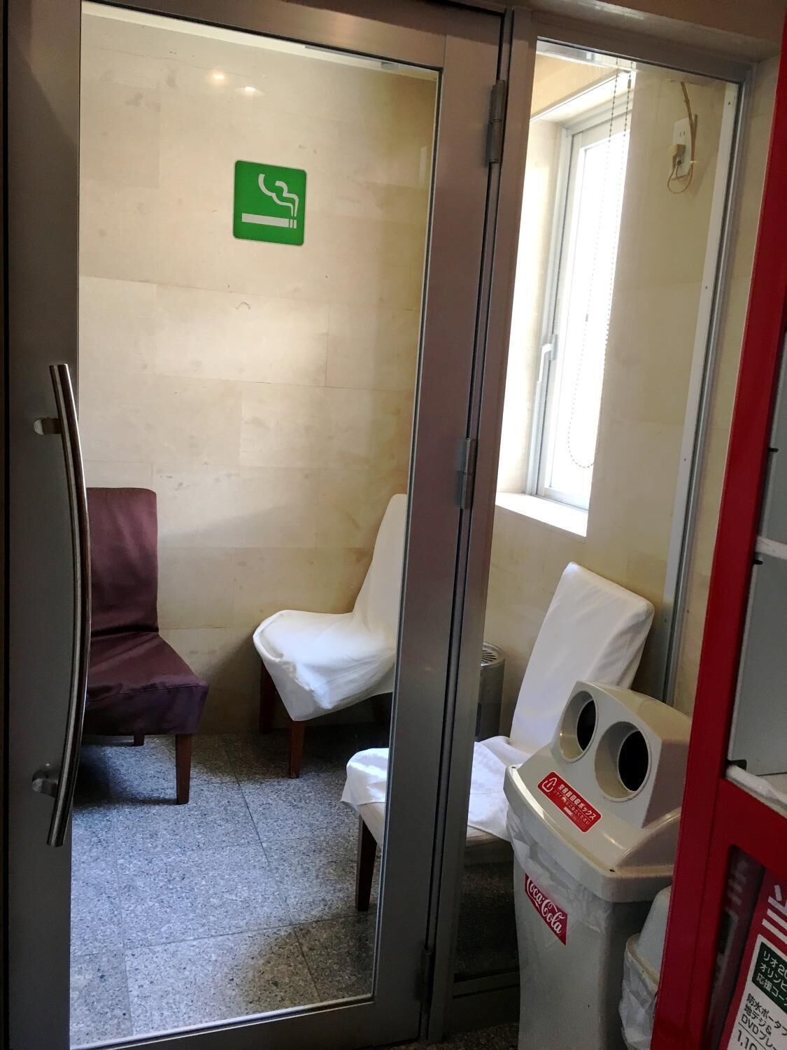 A restroom with a glass partition, a green emergency exit sign, a Coca-Cola trash bin, and two chairs.