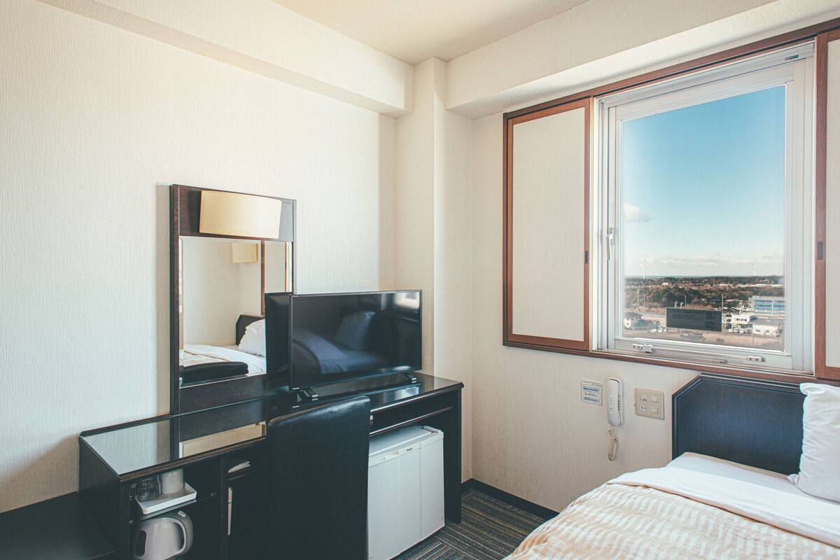 Una camera d'hotel con un letto, una scrivania con un computer, uno specchio, una finestra con vista sulla città e un telefono.