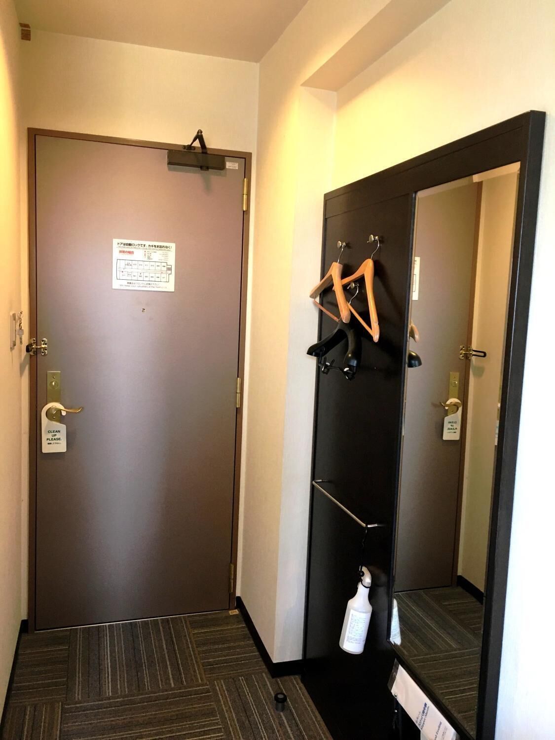 A hotel room with a closed door, a coat rack with hangers, a small table with a bottle, and a mirror reflecting the room.
