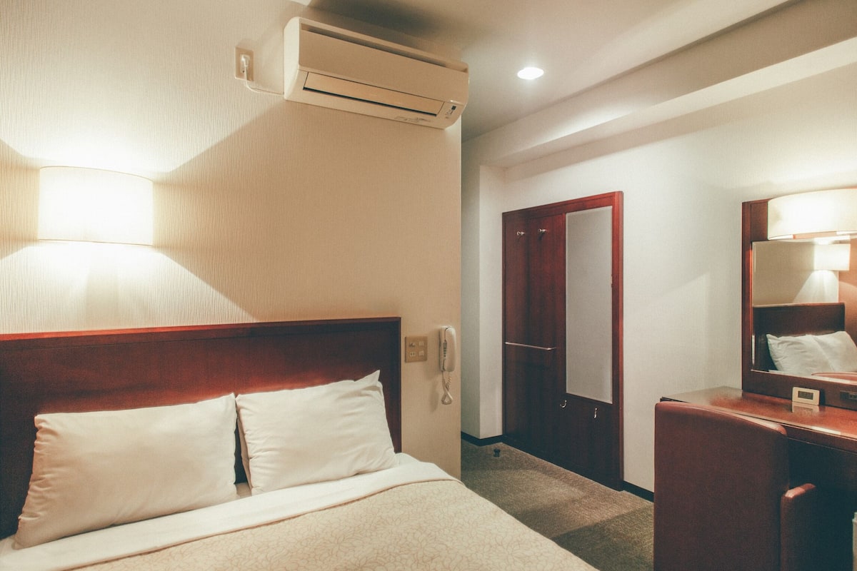 A hotel room with a bed, bedside lamp, air conditioning unit, wooden headboard, a mirror, and a desk with a lamp.