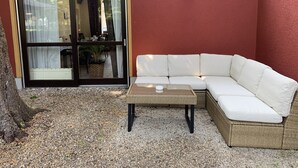 Un espacio exterior con mobiliario blanco, que incluye una mesa de mimbre y un sofá, una puerta corrediza de vidrio y una lámpara fijada a la pared.