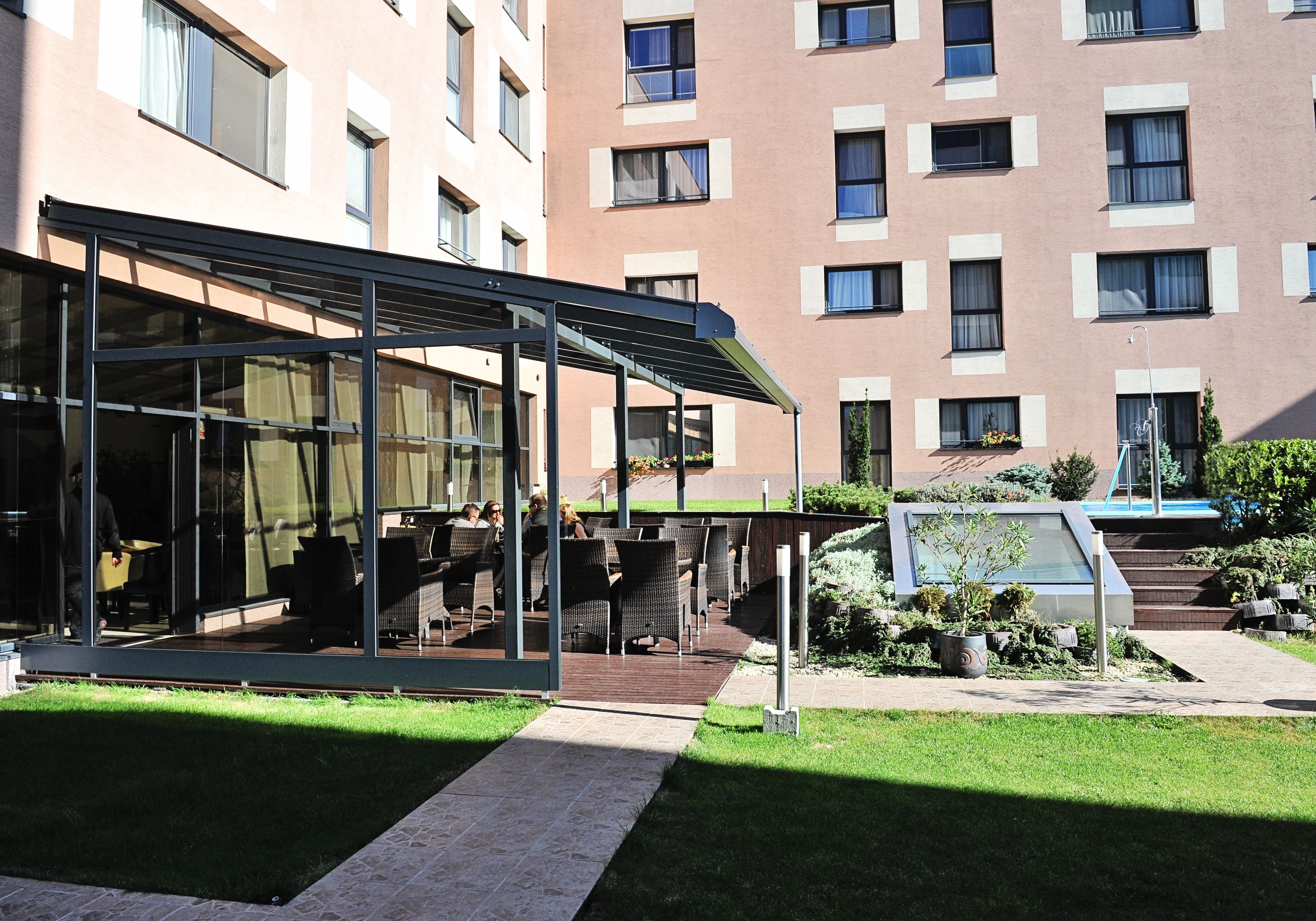 Un espace extérieur aménagé avec des murs en verre, une terrasse couverte et une piscine en arrière-plan.