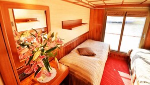 A cabin with a bed, a bedside table with a vase of flowers, a mirror, and a window with a view of the water.