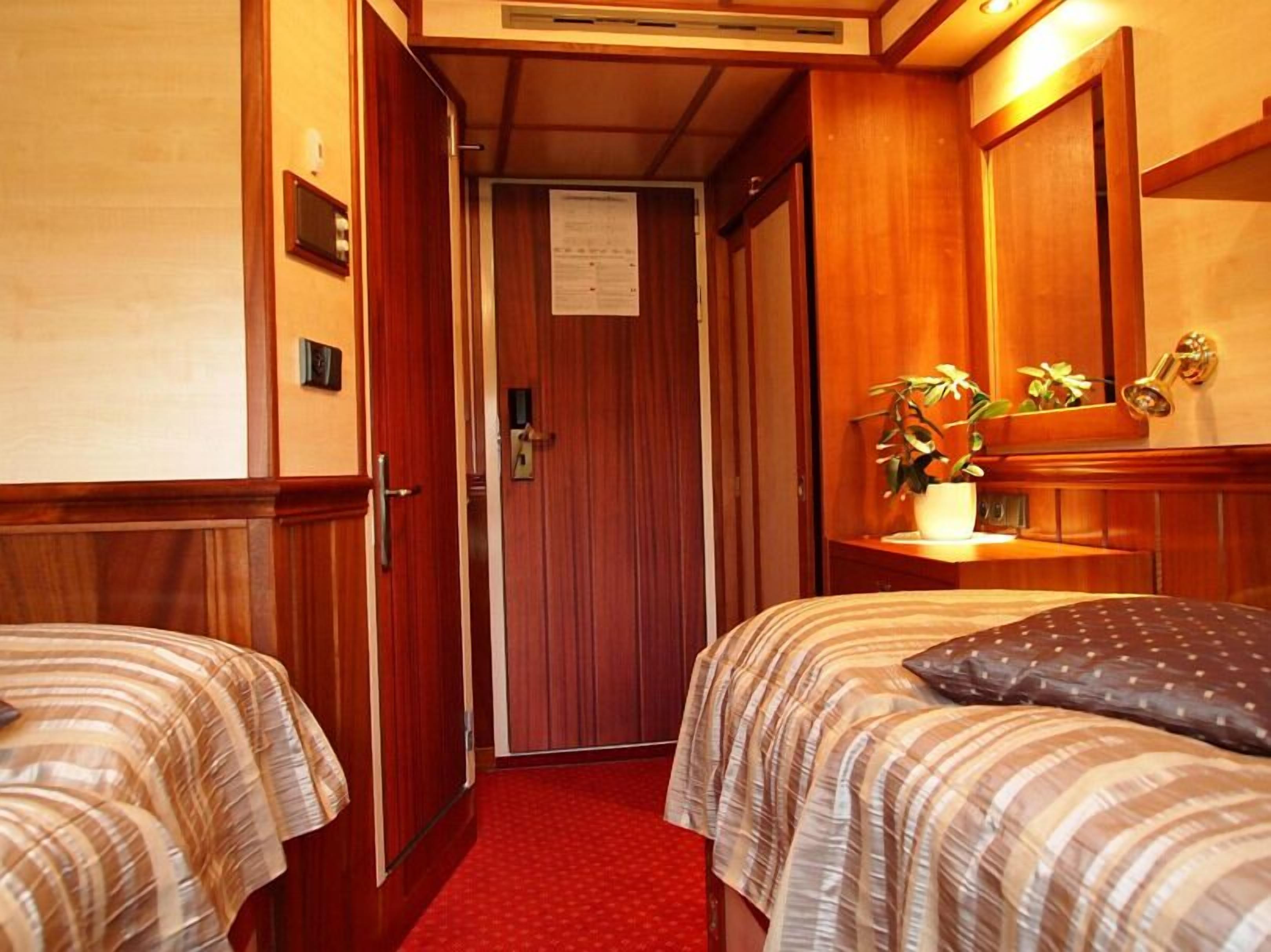 A narrow hallway with wooden walls and doors, leading to a room with two beds and a small table with a plant.