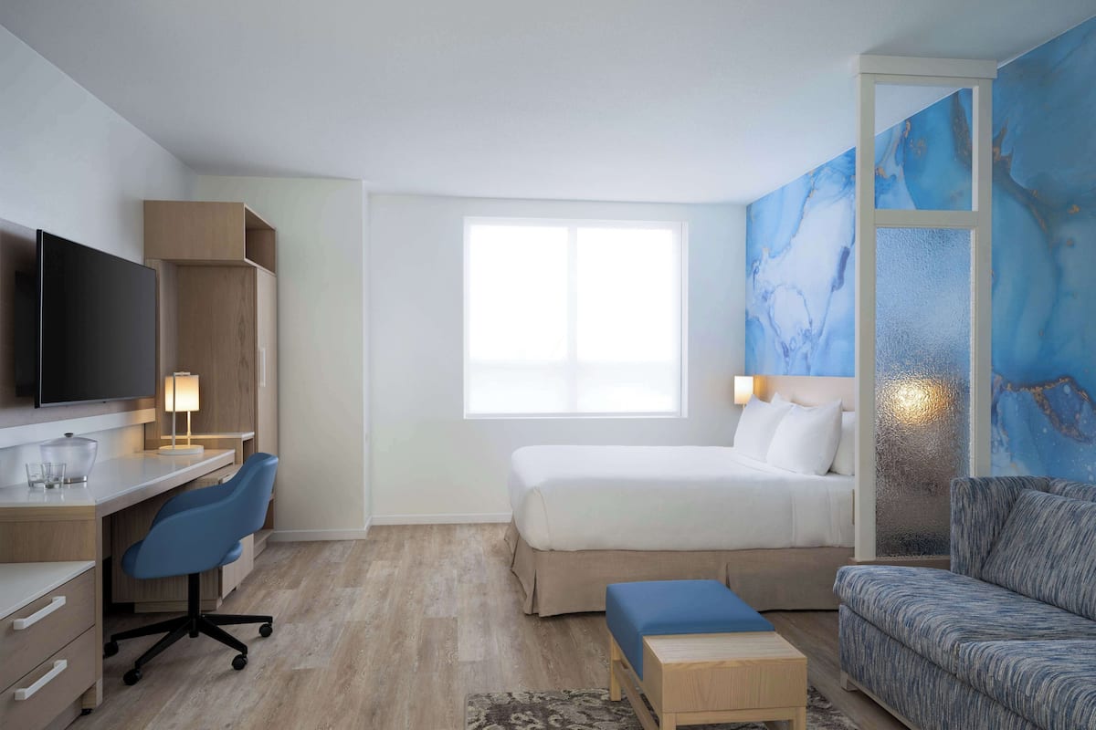Ett modernt hotellrum med en säng, ett skrivbord med en stol, en soffa och ett stort fönster.