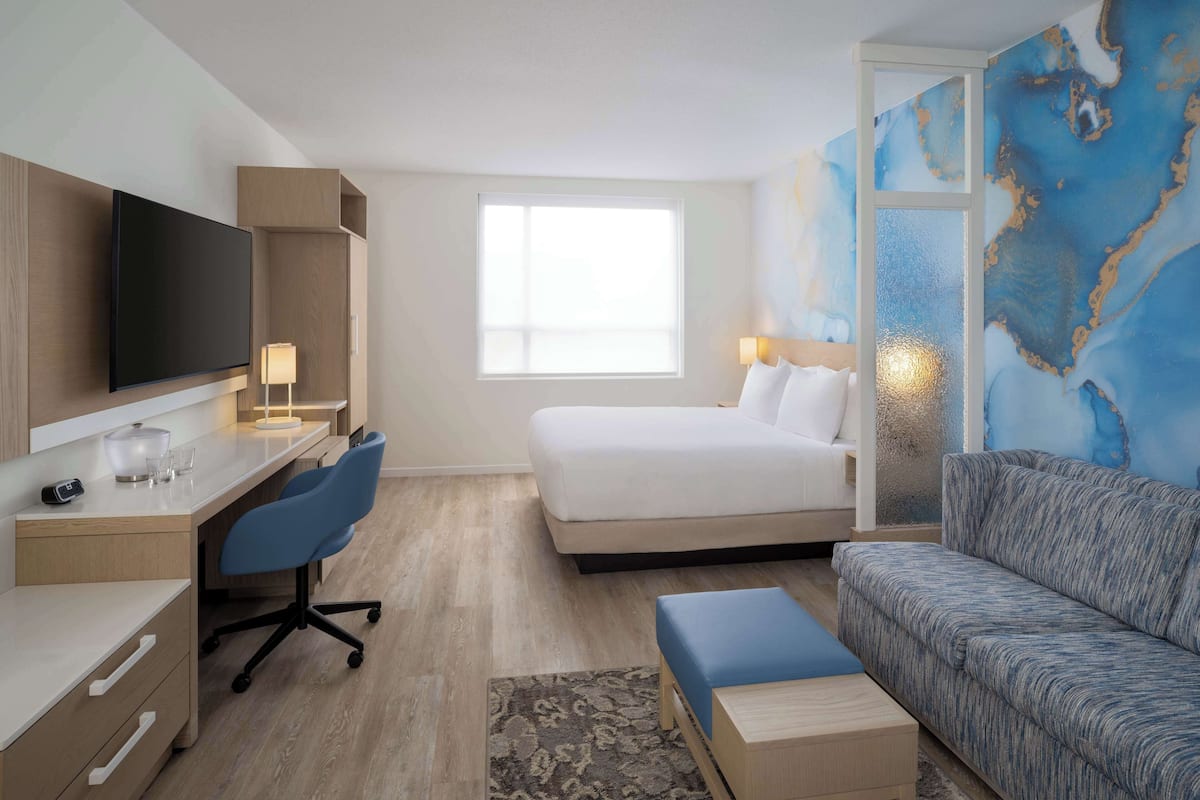 Ett hotellrum med en säng, ett skrivbord med en stol, en tv och ett stort fönster.