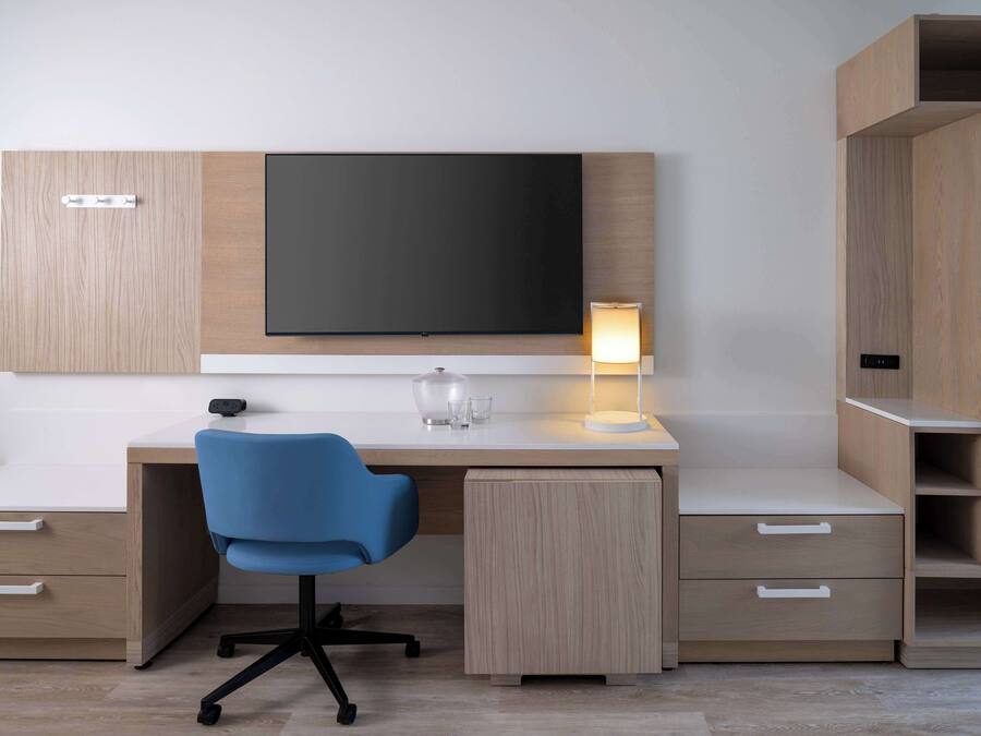 Una habitación de hotel moderna con un escritorio de madera, una silla de oficina azul, un televisor de pantalla plana en la pared, una lámpara de noche y estanterías empotradas.