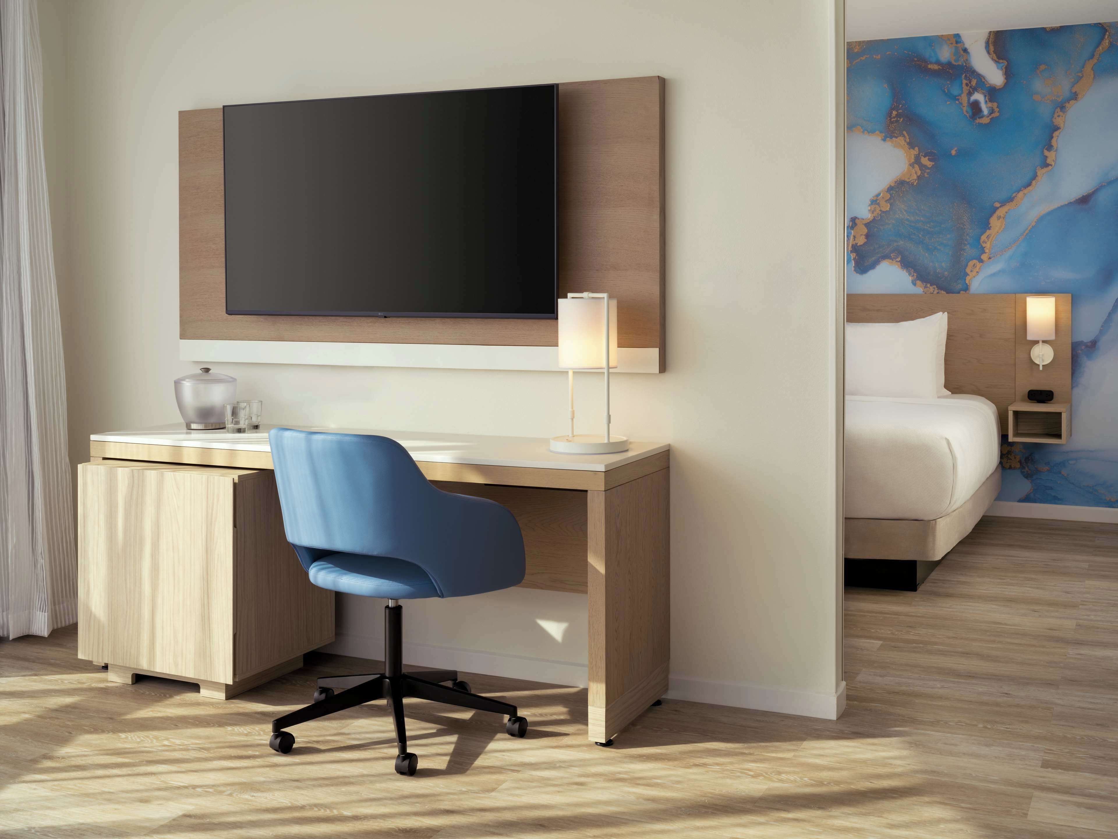 Ein Hotelzimmer mit einem Holztisch, einem blauen Sessel, einem Bett, einem Fernseher und einer Wand mit blauem abstrakten Design.