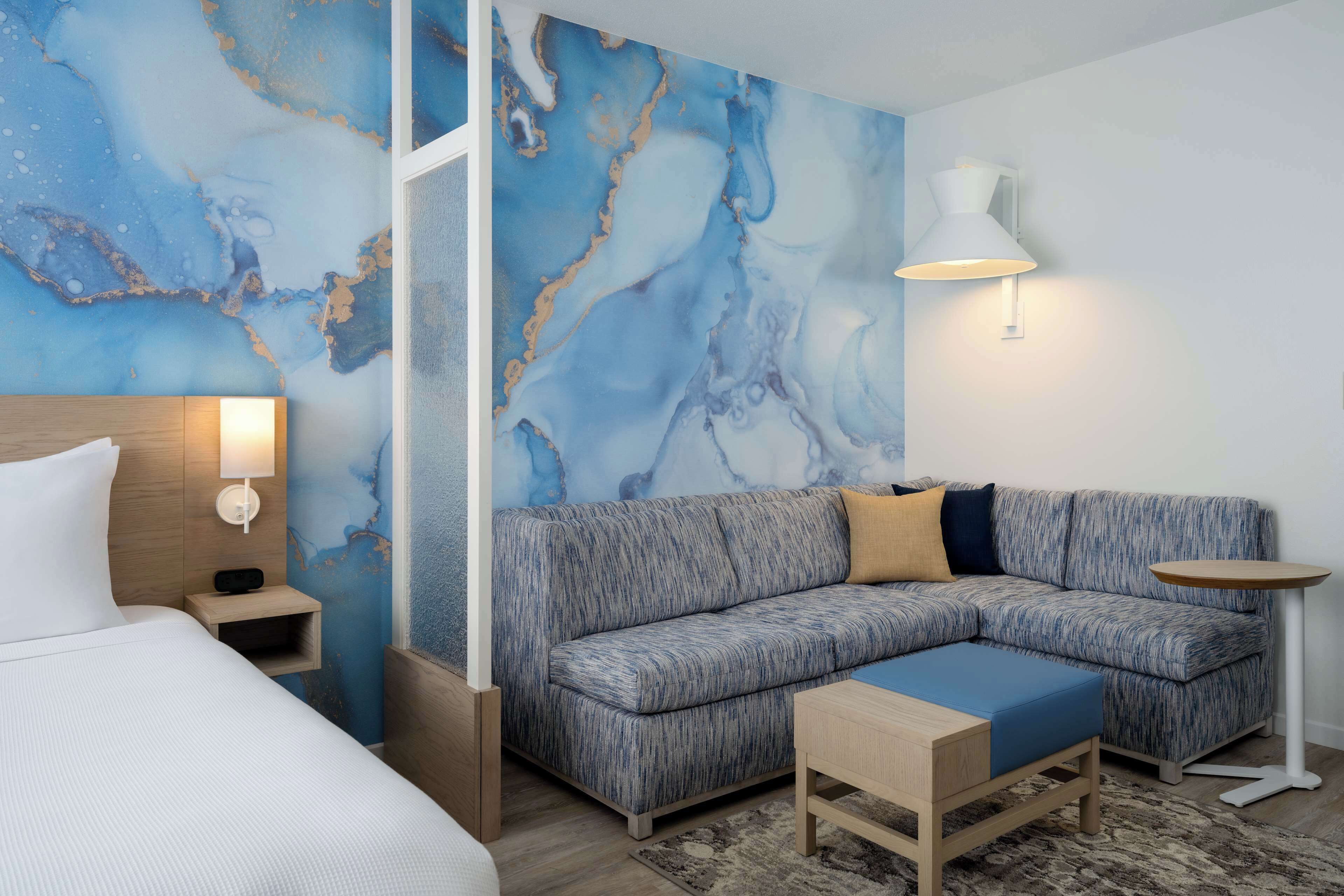 A modern hotel room with a sectional sofa, a small wooden table, a bedside lamp, and a wall with a blue marble design.