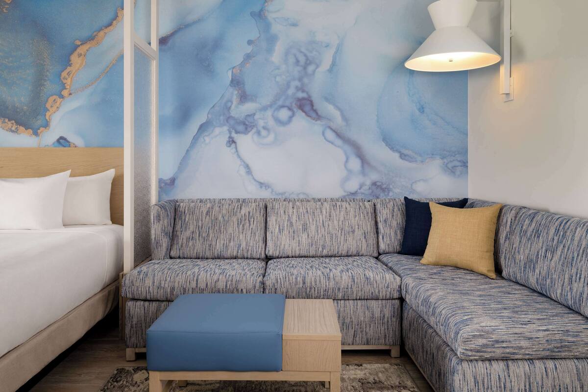 Ett modernt hotellrum med en stor, blå, abstrakt väggmålning, en grå klädd soffa, ett träbord som sängbord och en vit sänglampa.