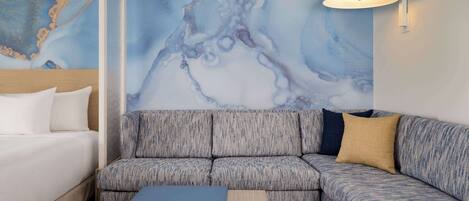 A modern hotel room with a large blue abstract mural, a grey upholstered sofa, a wooden nightstand, and a white bedside lamp.