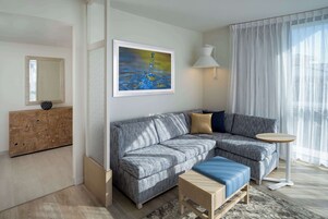 A modern living room with a grey sectional sofa, a small wooden coffee table, a round side table, a large mirror, and a painting on the wall.