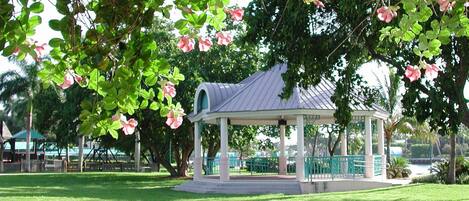 A gazebo with a green roof and white columns, surrounded by lush green grass and pink flowers.