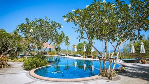 Una piscina circular rodeada de sillones de descanso y sombrillas, con árboles y un cielo azul despejado al fondo.