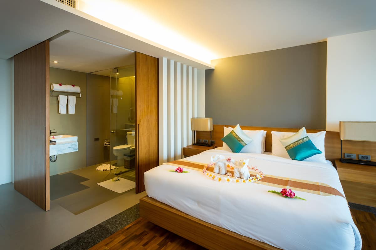 A modern hotel room with a large bed, a wooden headboard, a bedside table with a lamp, a glass shower enclosure, and a wooden wardrobe.