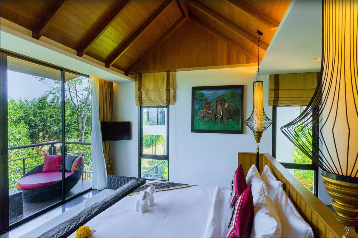 A room with a large bed, a window with a view of greenery, a painting on the wall, and a unique lamp.