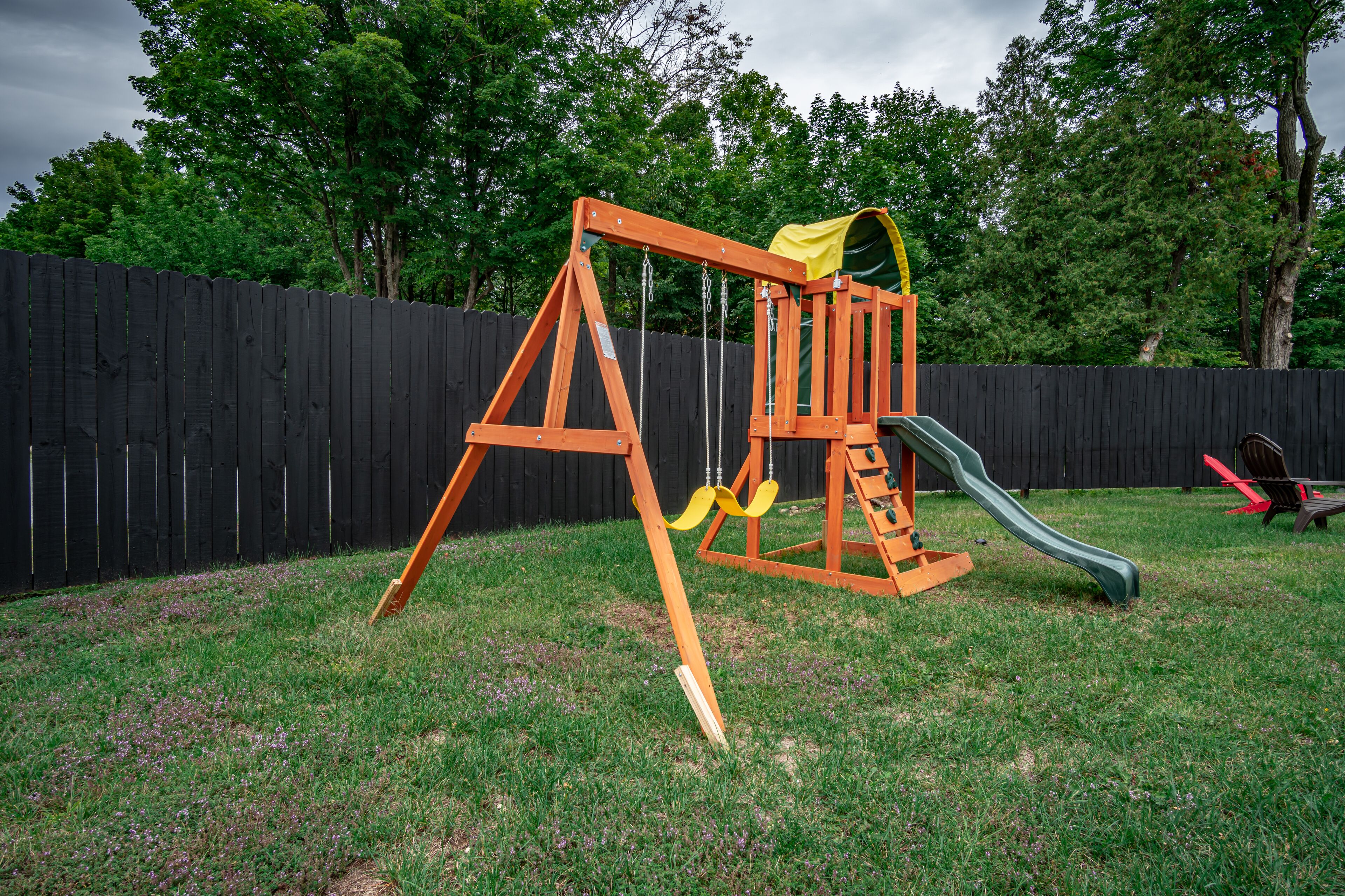 A wooden swing set with a slide in a backyard with a wooden fence and trees in the background.