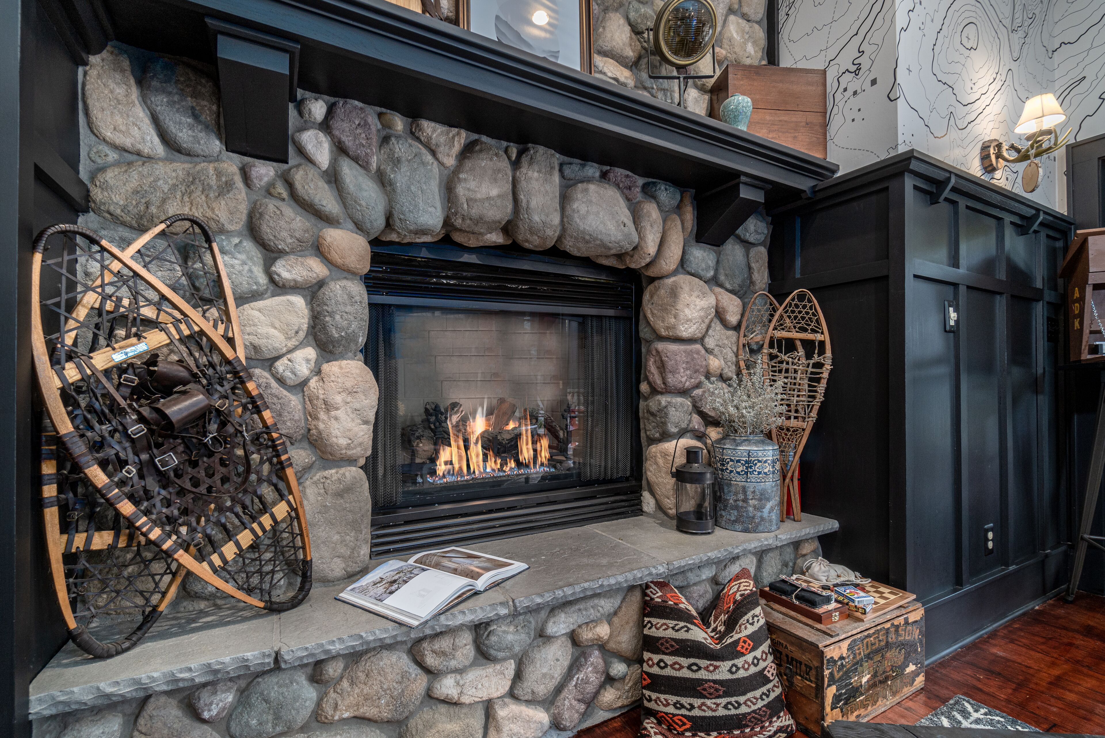 A stone fireplace with a fire, surrounded by rustic decor including a woven basket, a lantern, and a patterned cushion.