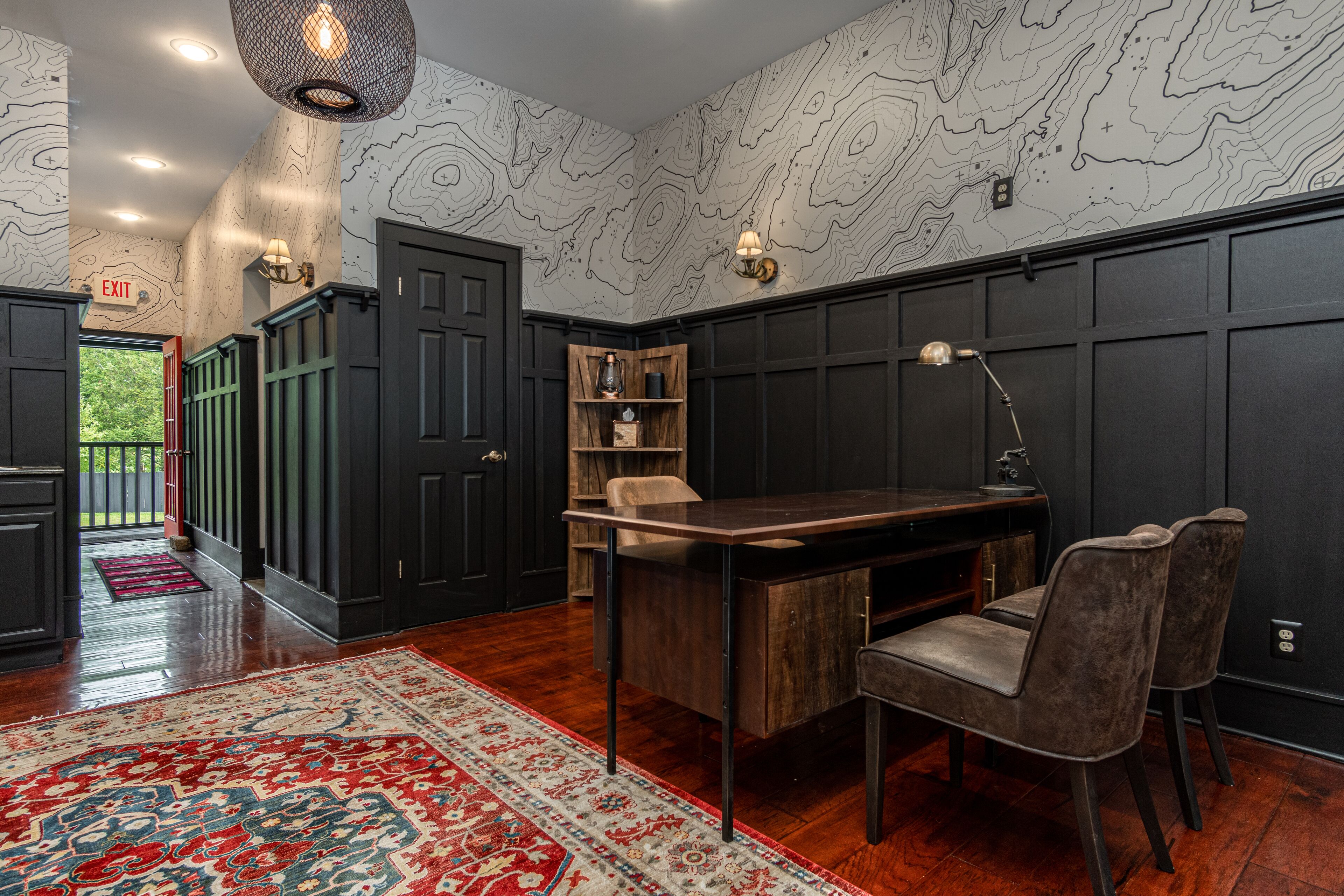 A modern interior with a dark wood dining table, two chairs, a red and white patterned rug, and a wall with a topographic design.