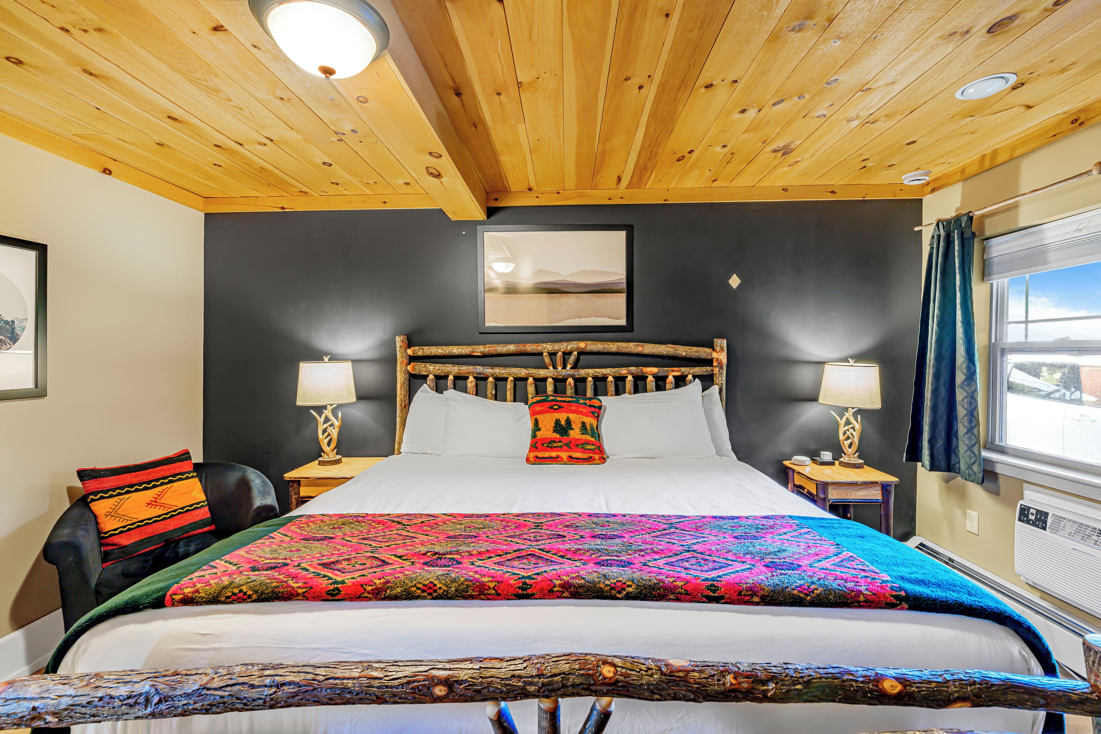 A room with a wooden ceiling, a bed with a colorful blanket, a headboard with a decorative pillow, a nightstand with a lamp, and a window with curtains.
