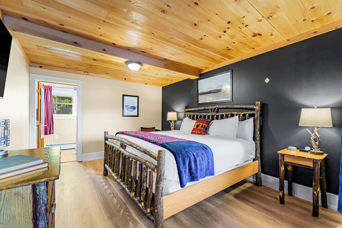 A bedroom with a wooden ceiling, a bed with a headboard, a nightstand, and a door with a curtain.