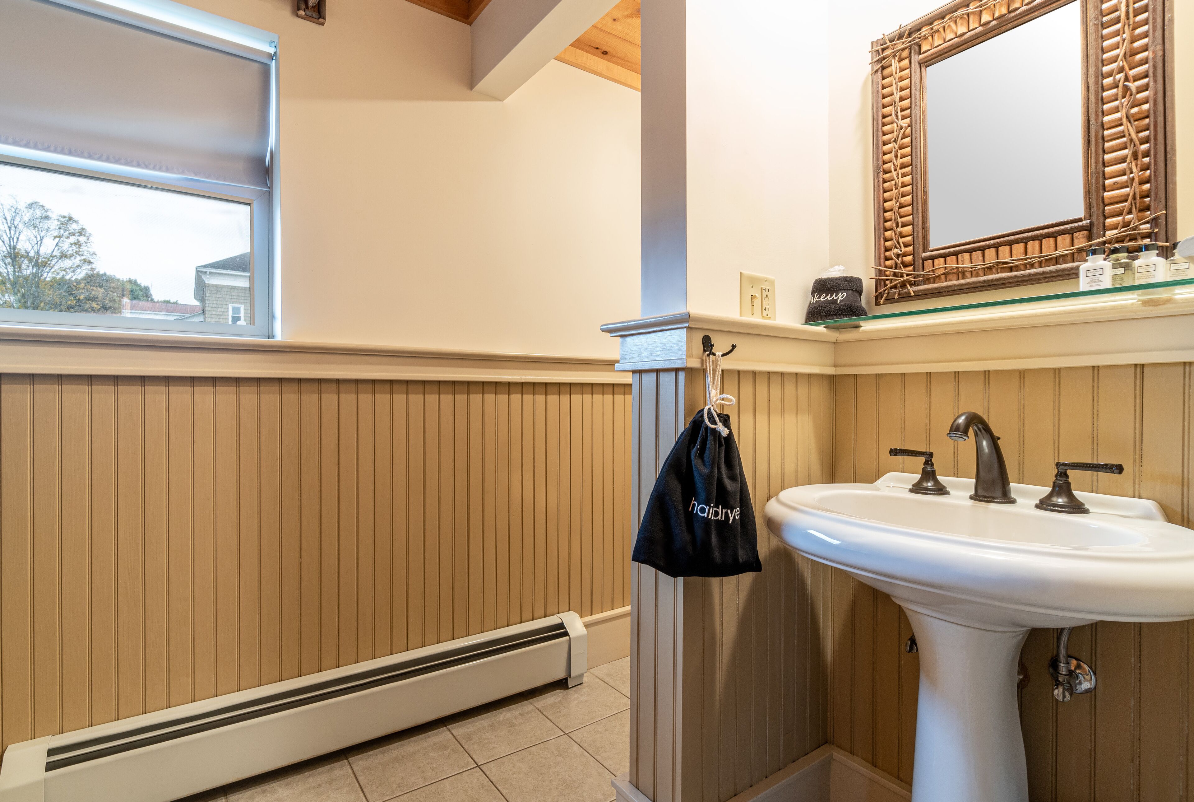 Une salle de bain avec un lavabo blanc, un lambris en bois, un miroir avec un cadre tressé et une fenêtre avec un store.