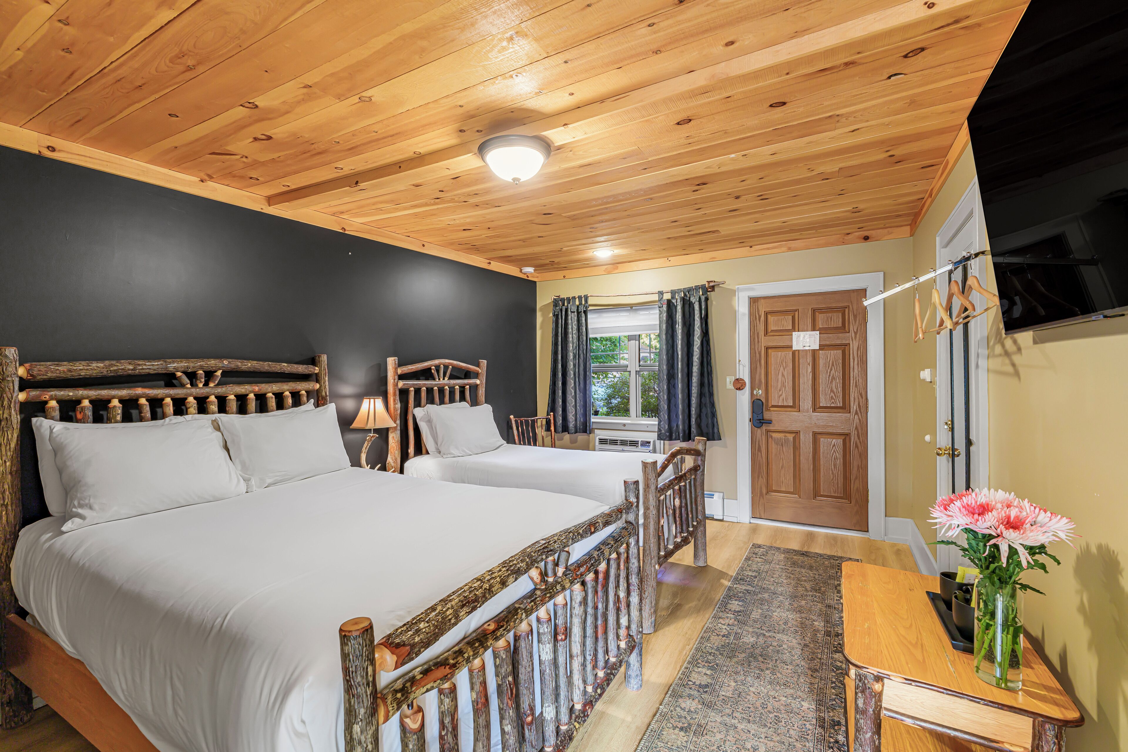 A room with two wooden beds, a wooden ceiling, a small table with a vase of flowers, and a door with a window.