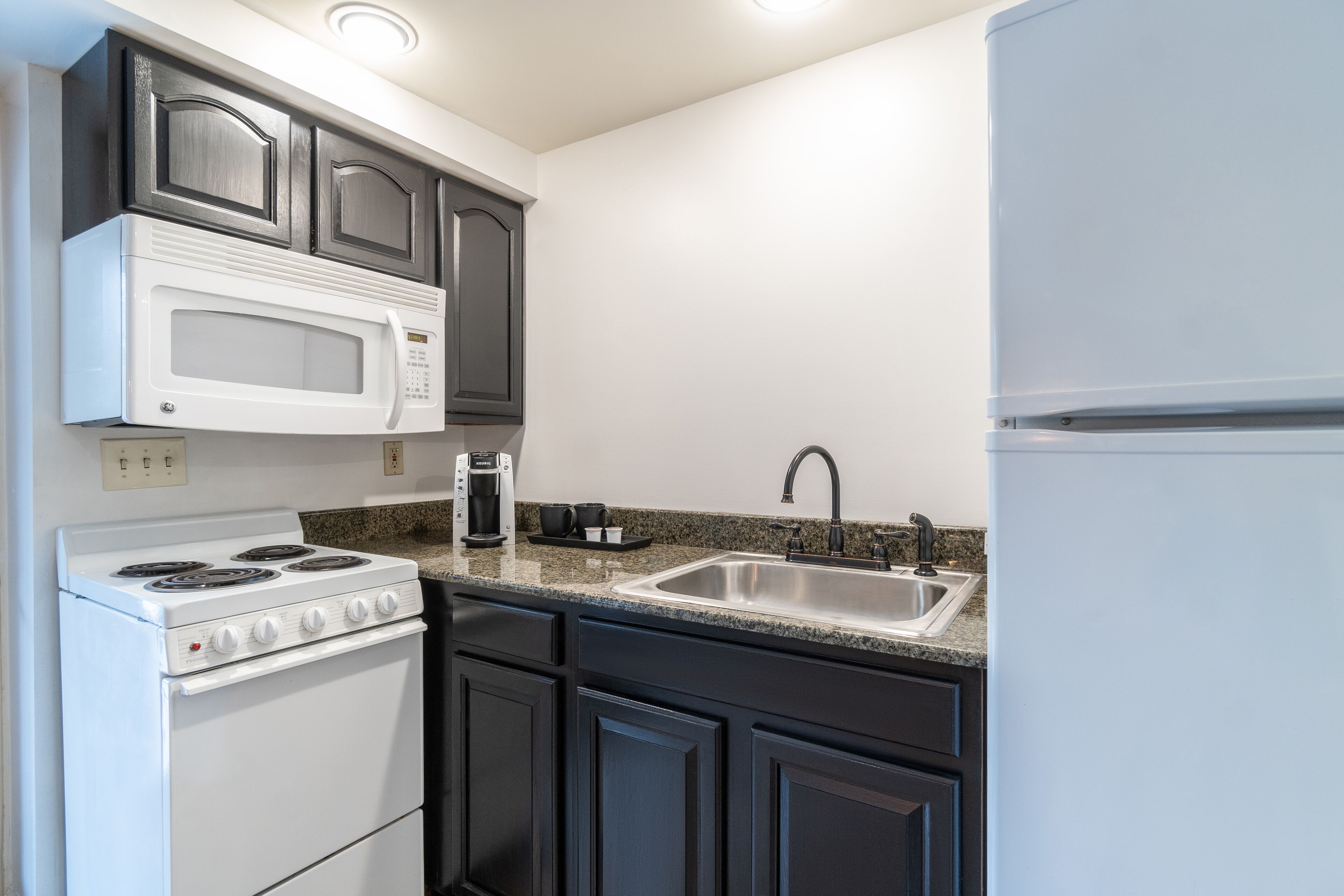 A kitchen with dark cabinets, a white stove, a microwave, a sink, and a coffee maker.