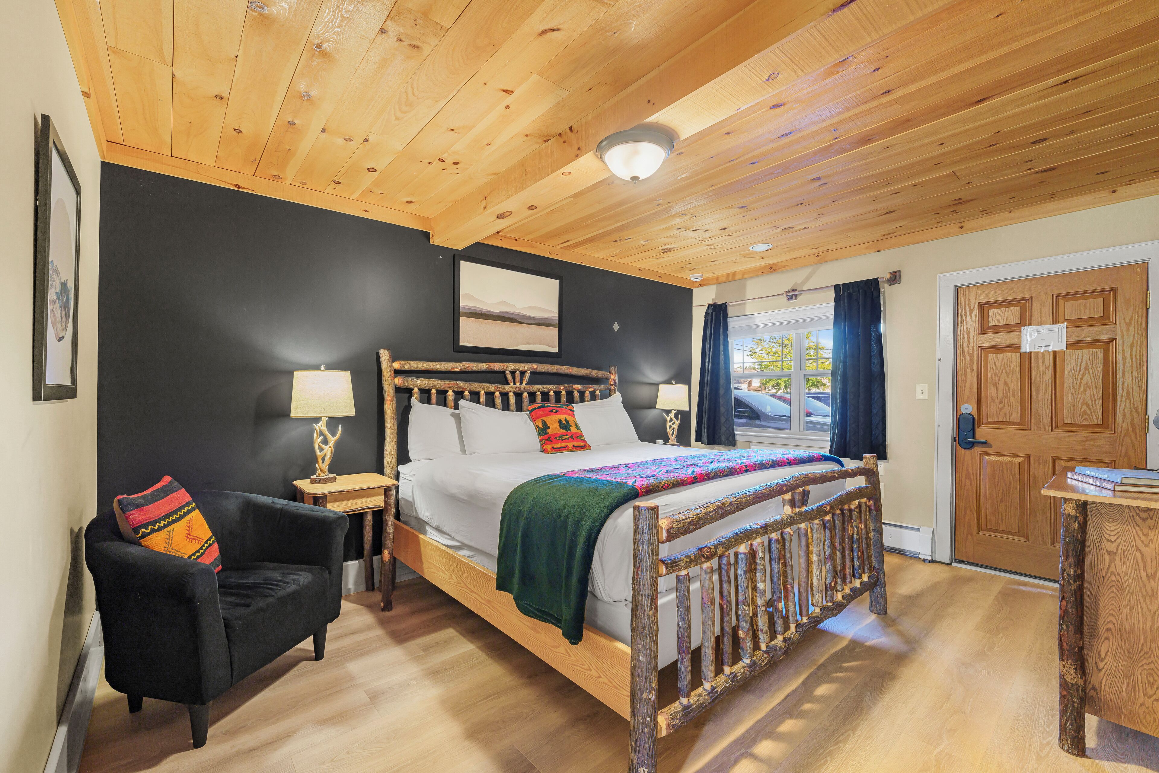 A wooden ceiling, a bed with pillows, a chair, a nightstand, a lamp, a door, and a window.