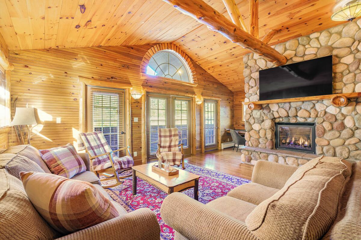 A cozy log cabin interior with a stone fireplace, wooden ceiling, and a large arched window.