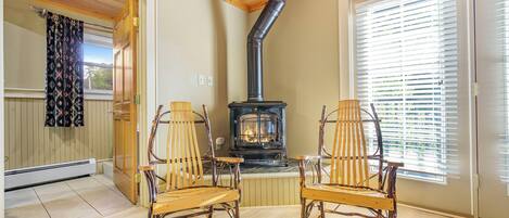 A cozy room with a wood-burning stove, two rocking chairs, and a large window with blinds.