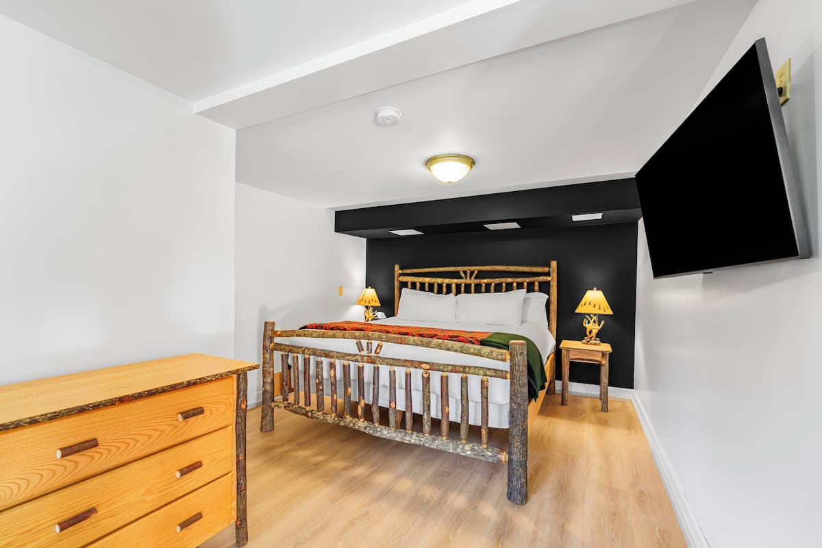 A bedroom with a wooden bed, a wooden dresser, a bedside table with a lamp, and a mounted TV.