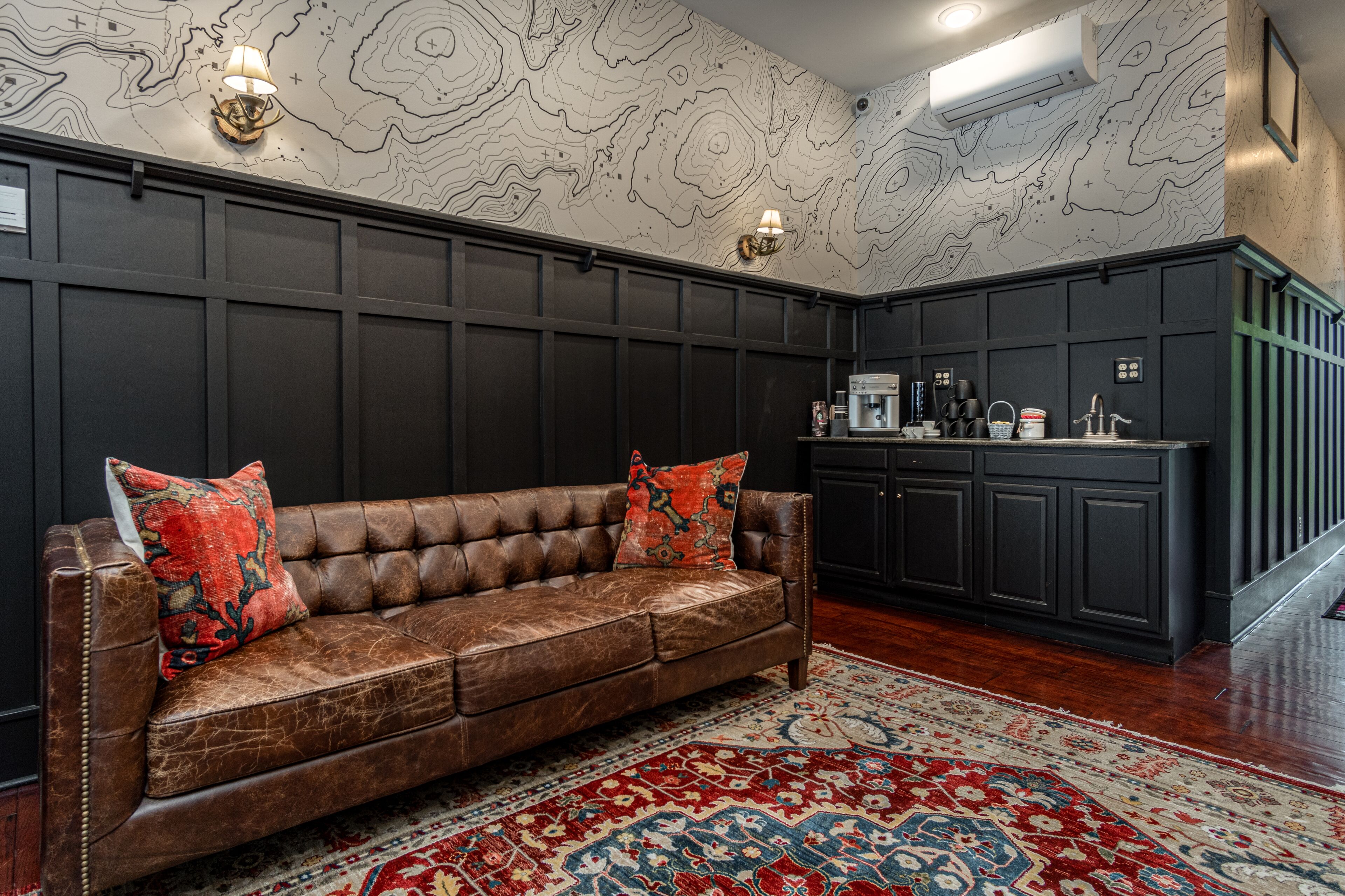 A brown leather sofa with red and black cushions, a patterned rug, and a kitchen area with dark cabinetry and a coffee machine.