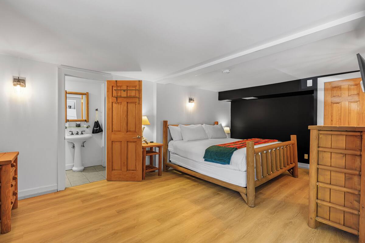 A bedroom with a wooden bed, a bedside table, a nightstand, a wardrobe, and a bathroom with a sink and mirror.