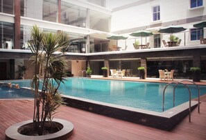A swimming pool with a wooden deck, surrounded by lounge chairs and umbrellas, and a glass balustrade above.